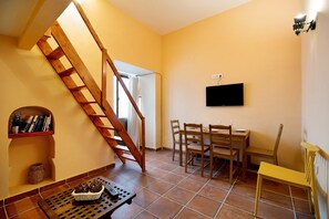 A room with a wooden staircase, a dining table with chairs, a TV mounted on the wall, and a shelf with books and decorative items.