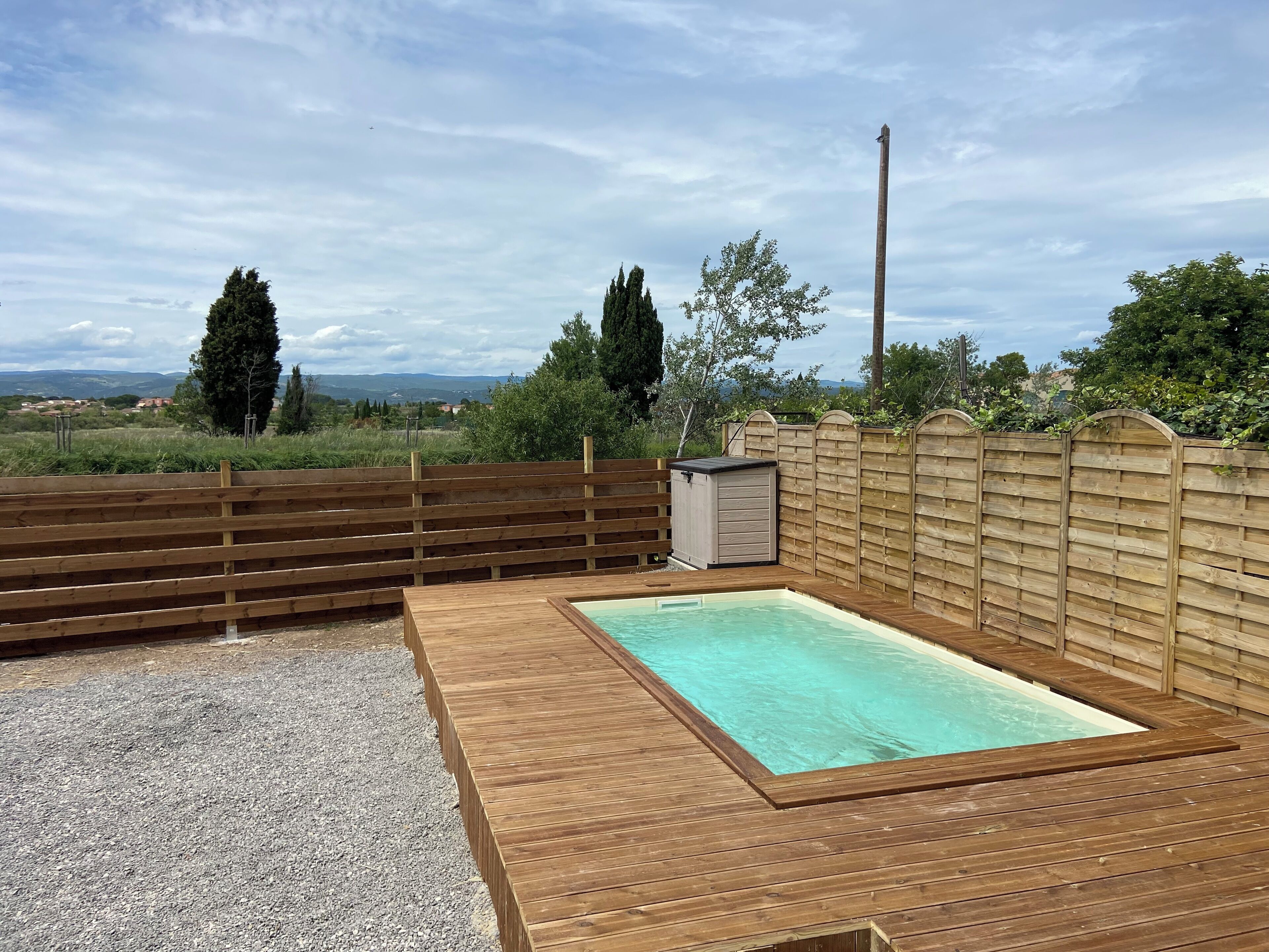 Una terrassa de fusta amb una piscina rectangular, envoltada per una tanca de fusta i un cobert petit.