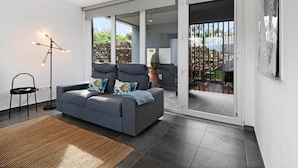 A modern living room with a grey sofa, a floor lamp, and a large sliding glass door leading to an outdoor area. - Holiday Home "Rocha do Mar 1" with Sea View, Shared Pool & Wi-Fi (Biscoitos)