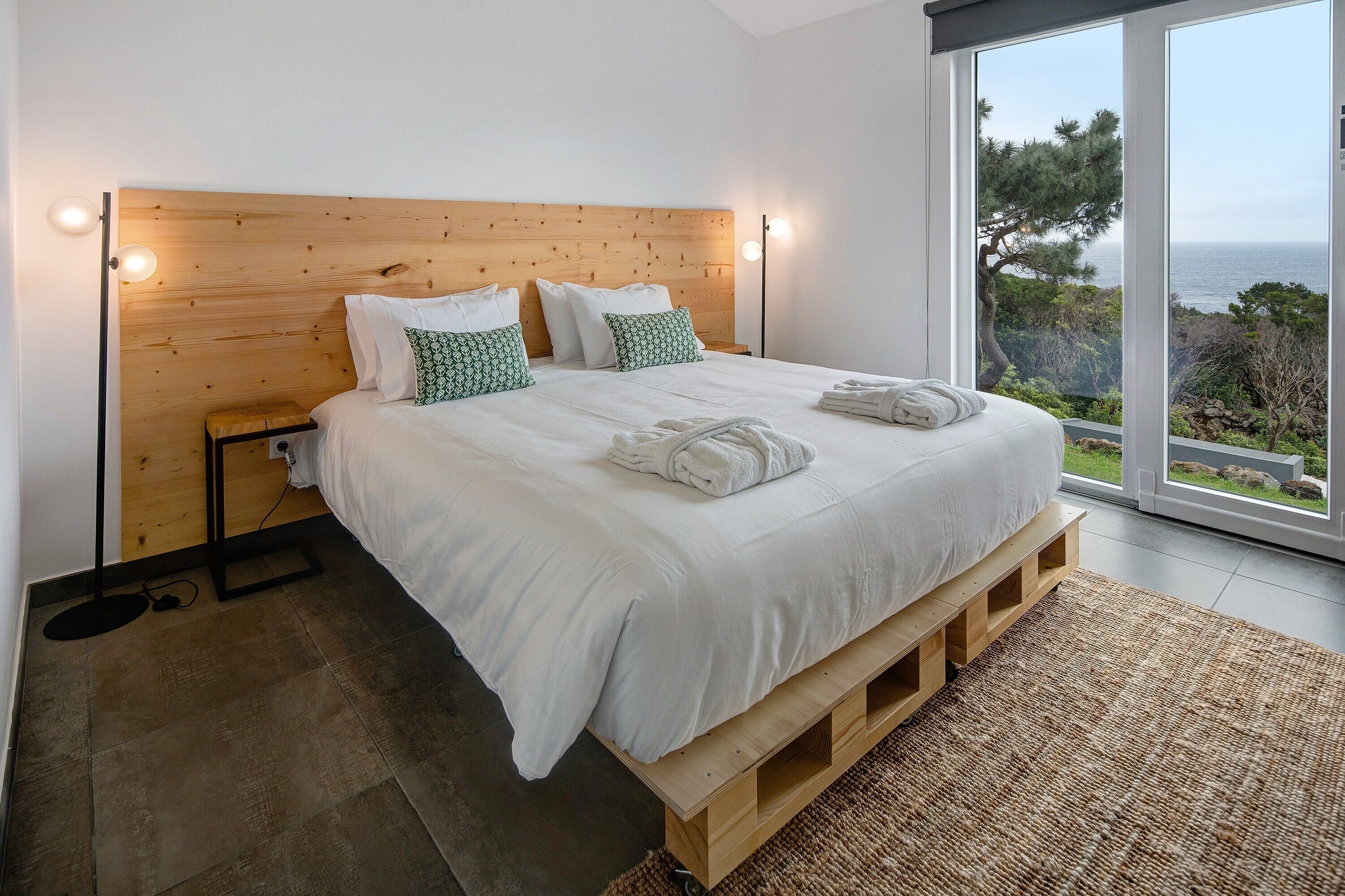 A bedroom with a wooden headboard, white bedding, a view of the sea, and a large window.