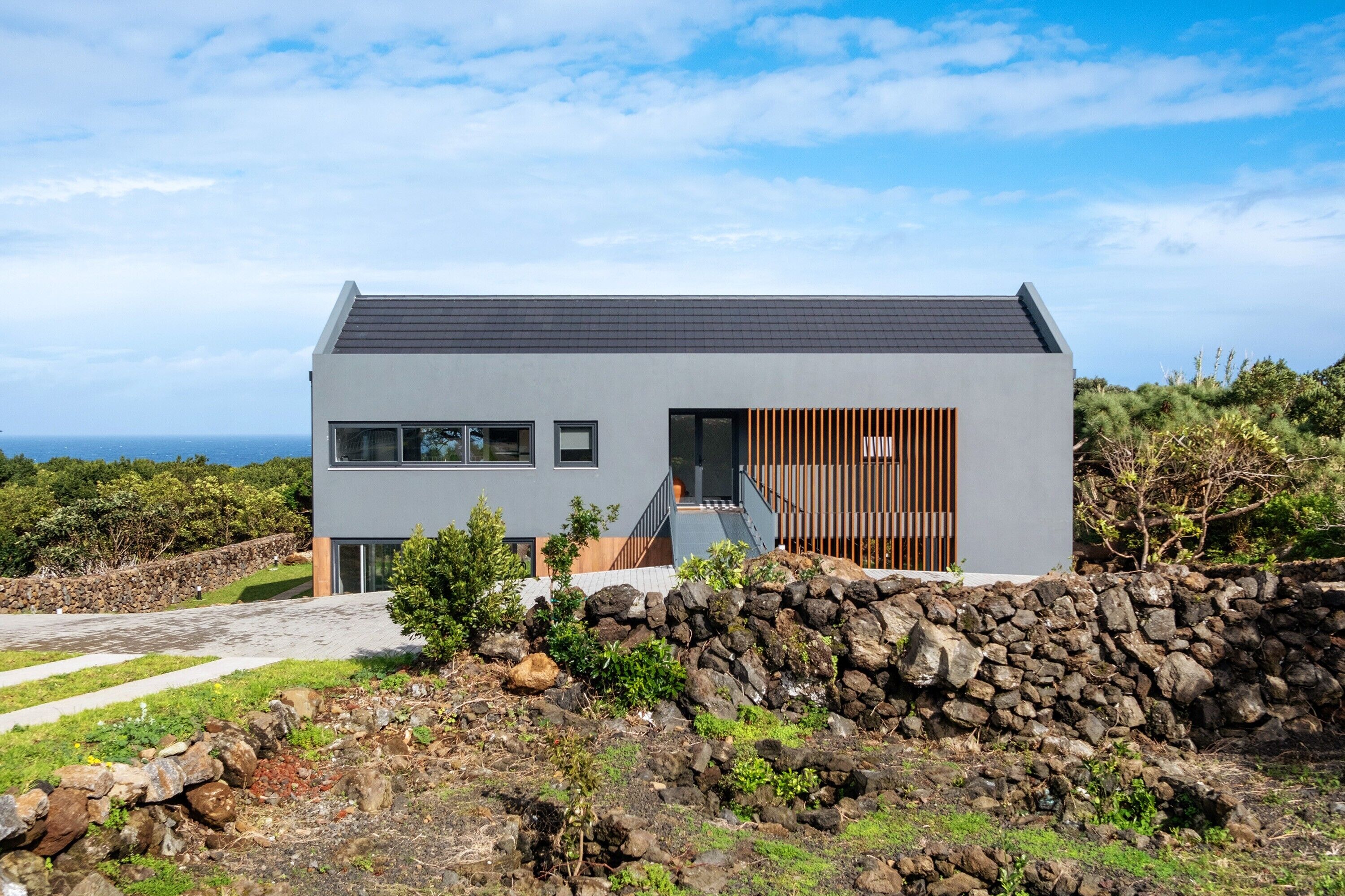 A modern house with a grey exterior, a stone wall in the foreground, and a view of the ocean.