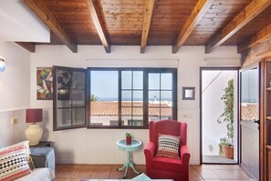 Una habitación con techo de madera, una silla roja, una pequeña mesa con una planta, una ventana con vistas, y una puerta que da al exterior.