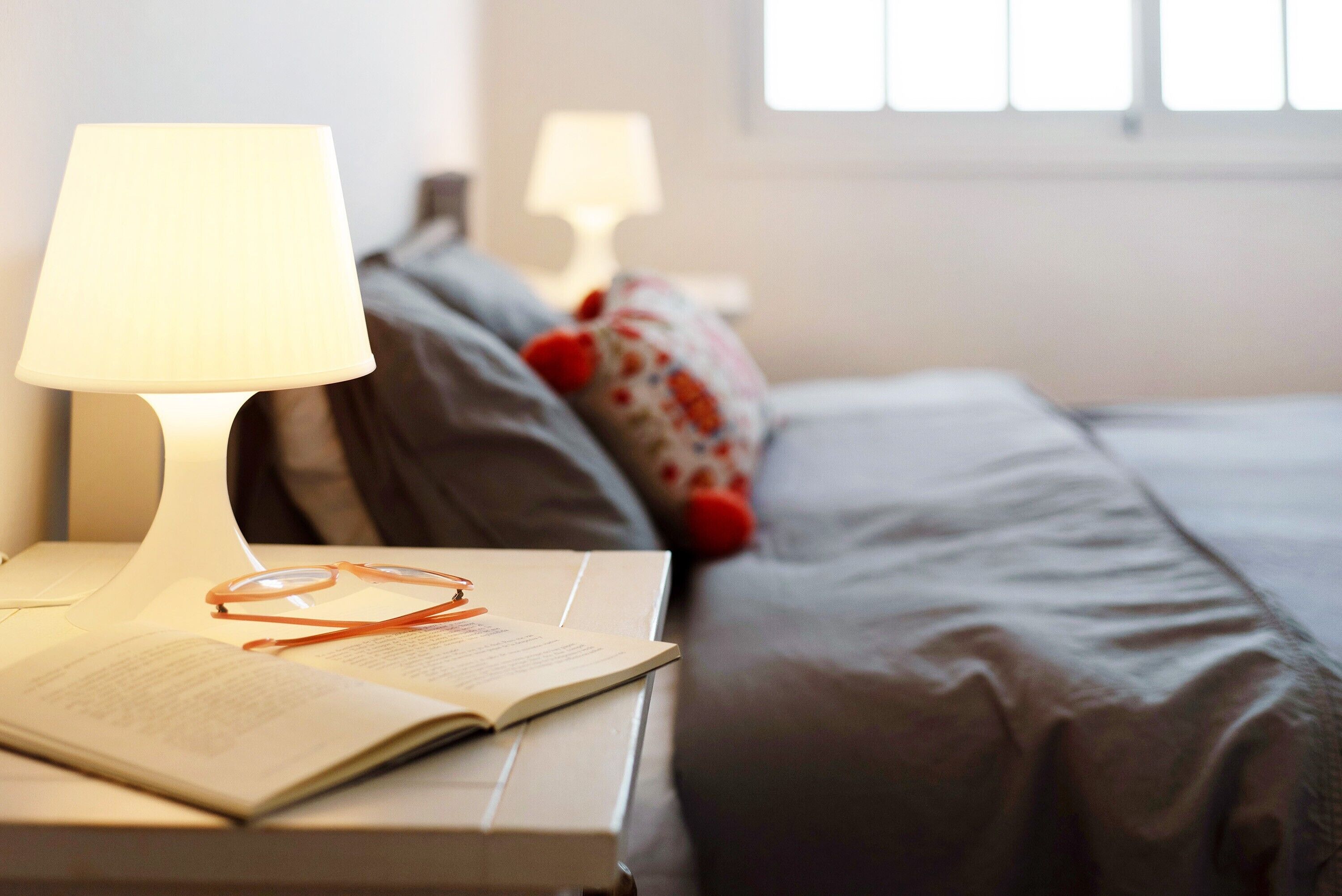 Una cama bien tendida con almohadas, una mesita de noche con lámpara, y un libro abierto con gafas encima.
