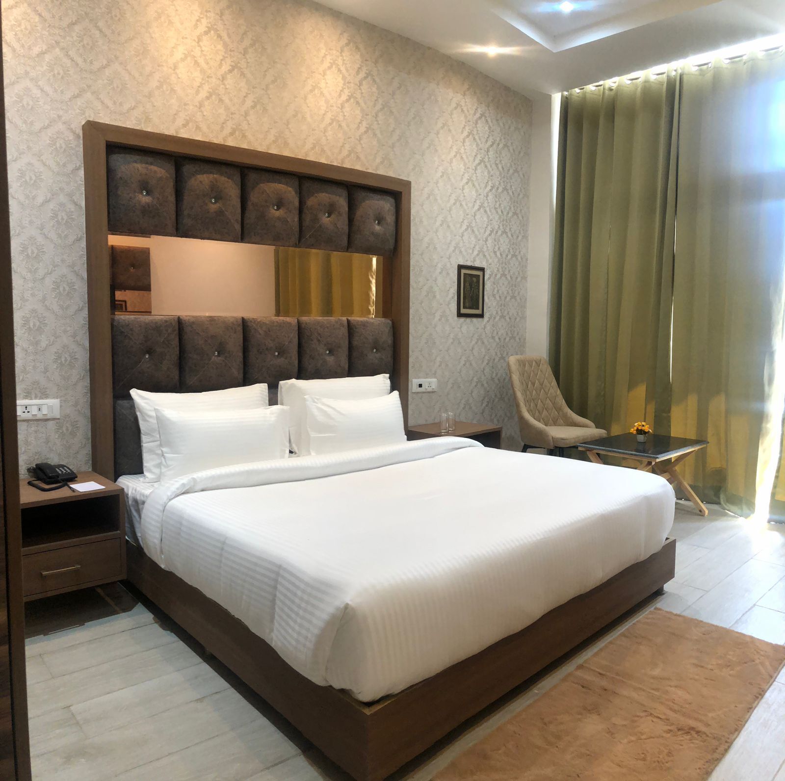 A large bed with a wooden headboard, a bedside table with a phone, and a chair with a small table beside it.