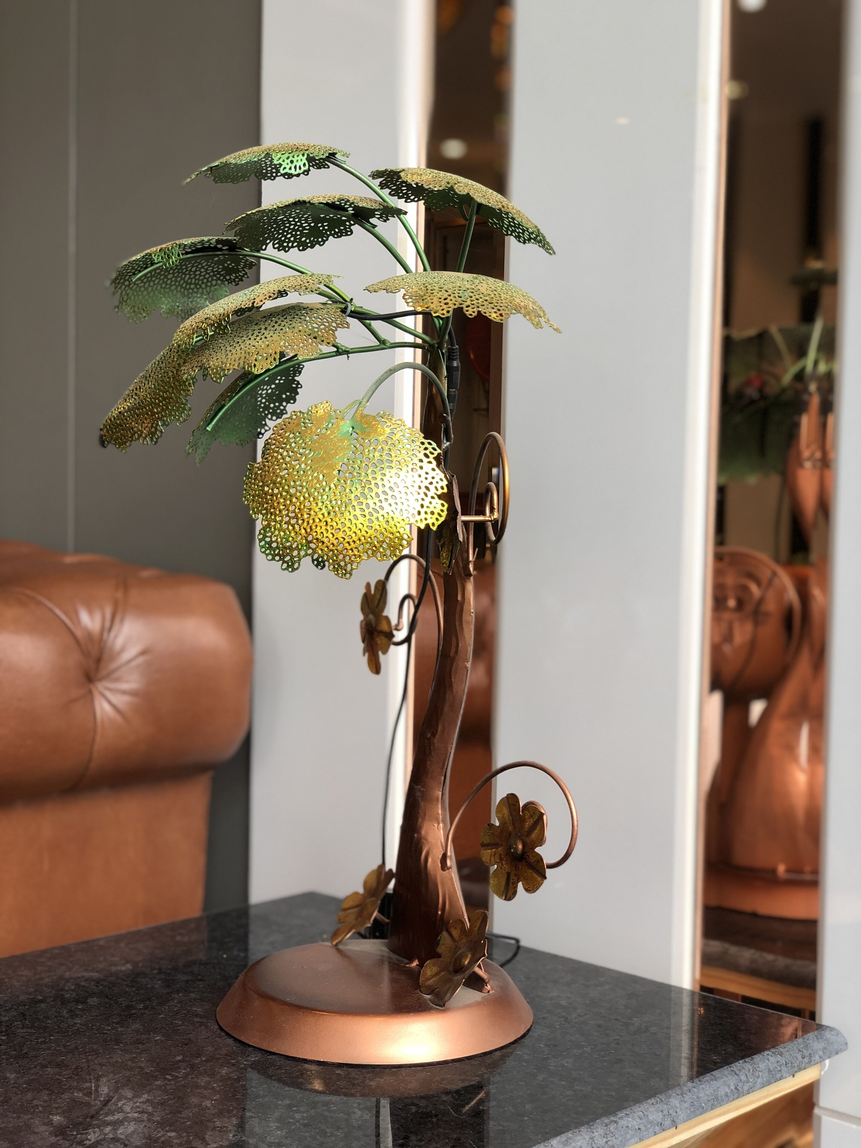 A copper tree sculpture with green leaves and a yellow flower on a black marble table.