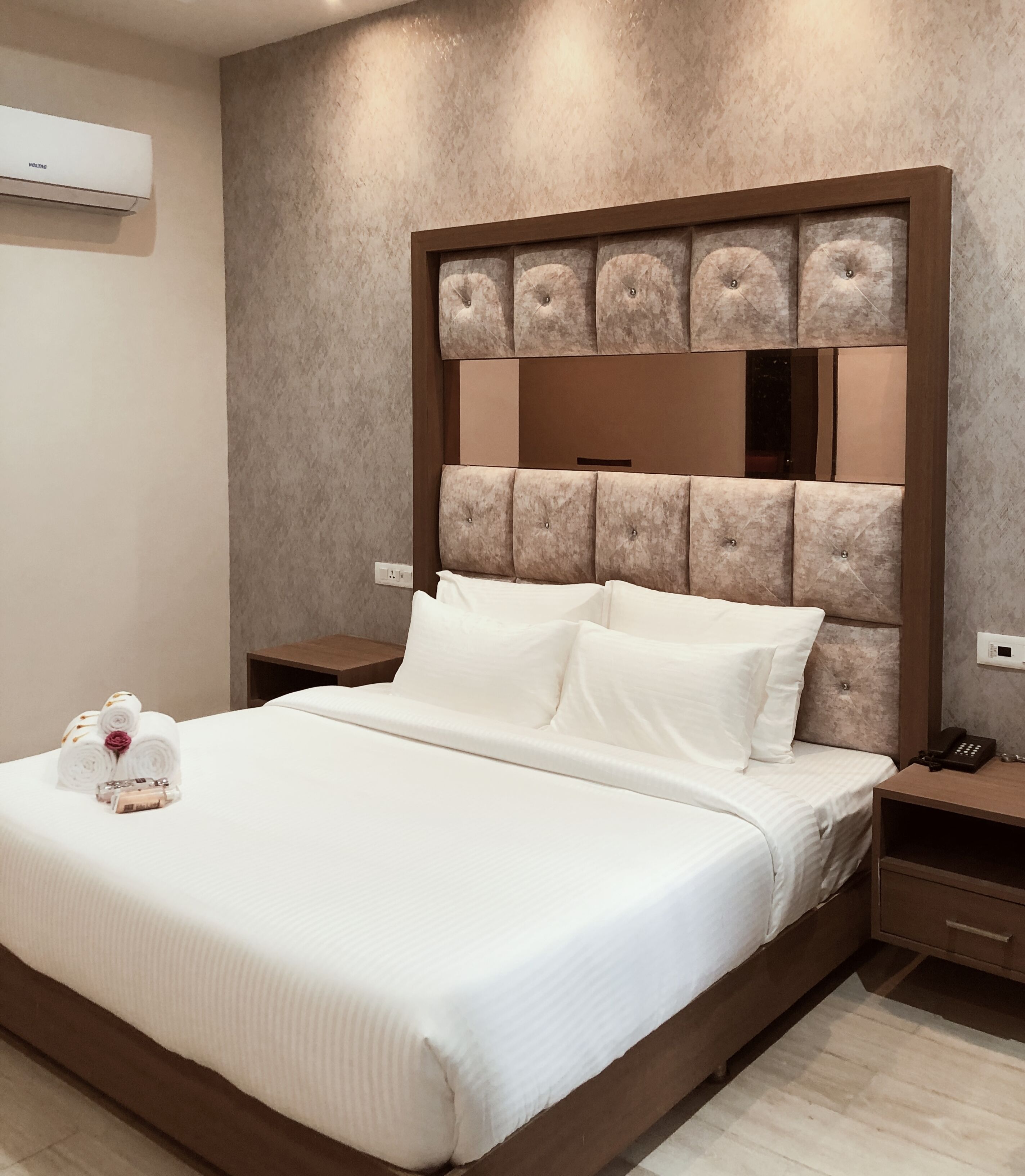 A hotel room with a large bed, a headboard with a mirror, a bedside table with a phone, and a wall-mounted air conditioner.