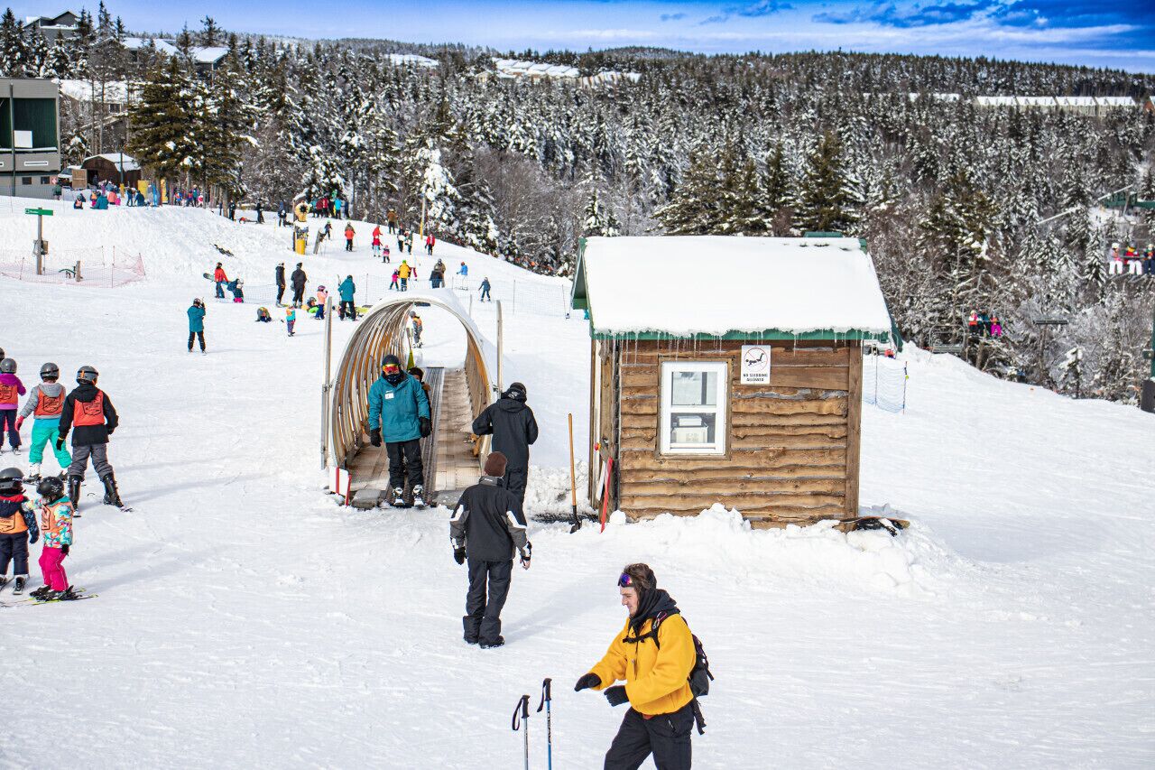 Une cabane en bois, dont le toit est recouvert de neige, située au pied d’une piste de ski.
