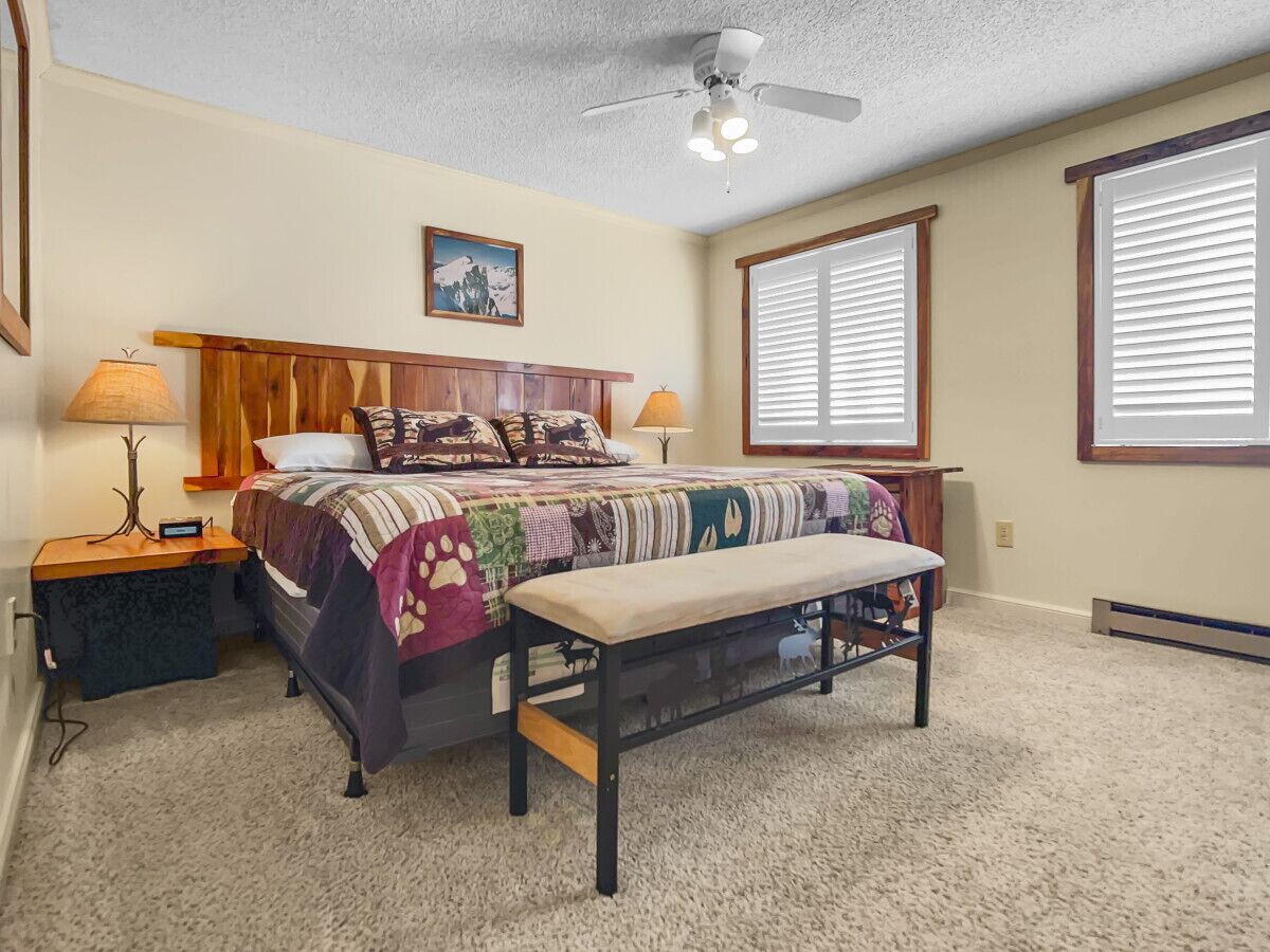 A bedroom with a bed, a bench, a nightstand, a lamp, a ceiling fan, and a window with blinds.