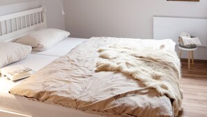 A bedroom with a white bed, wooden flooring, a small bedside table, and a window with natural light.