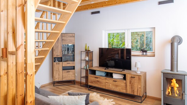 A modern living room with a wooden staircase, a TV, a fireplace, and a window with a view of greenery.