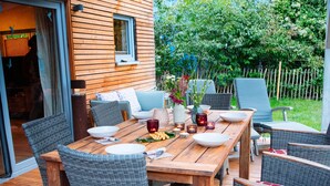 An outdoor dining area with a wooden table and wicker chairs, a covered seating area, and a view of a garden and fence.