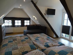 A loft bedroom with a sloped ceiling, a bed with patterned bedding, a bench, a TV mounted on the wall, and windows with blinds. - Fachwerkhaus in Ruhiger Umgebung in Direkter Nähe zum Dümmer See (Hüde)