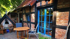 Un espace extérieur rustique aménagé avec des sièges, un bâtiment en briques, une table en bois et une fenêtre à cadre bleu.