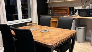 A dining area with a wooden table and black chairs, a kitchen with shelves of dishes and a microwave, and a window with a view of the sky. - Chez Caroline Ingalls (Gavray-sur-Sienne)