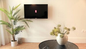 Een woonkamer met een flatscreen televisie aan de muur, een potplant, een klein rond tafeltje met een vaas en een afstandsbediening op tafel.