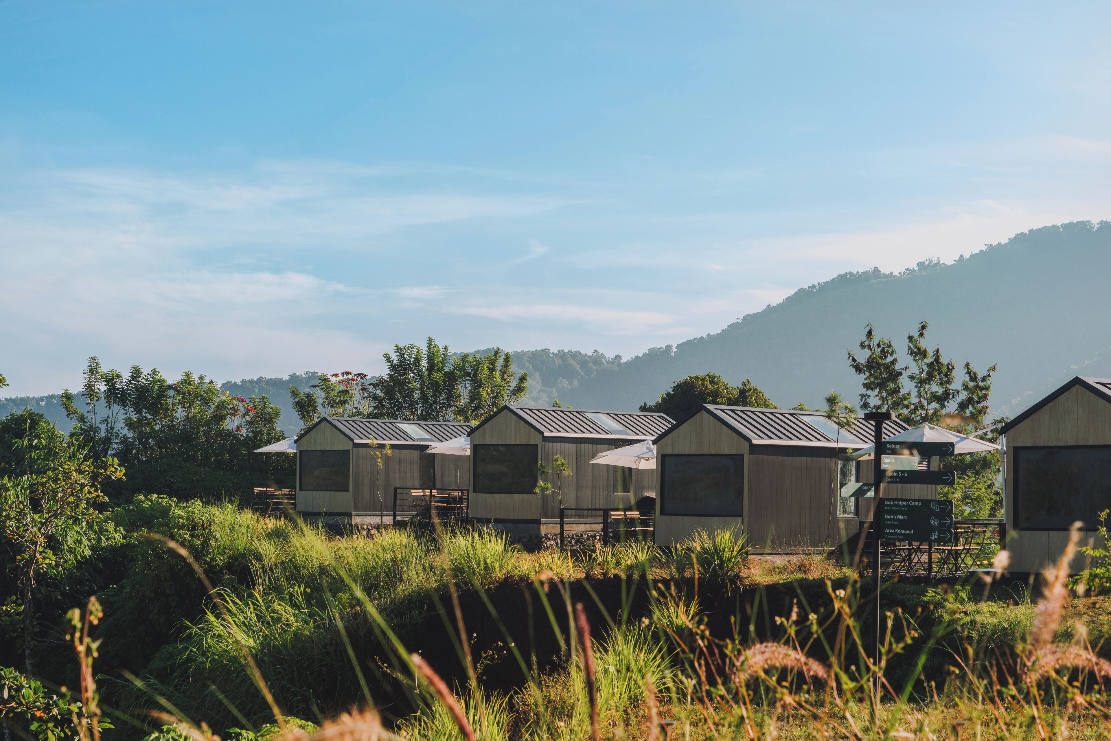 A row of small, single-story cabins with metal roofs, surrounded by lush greenery and a hilly backdrop.