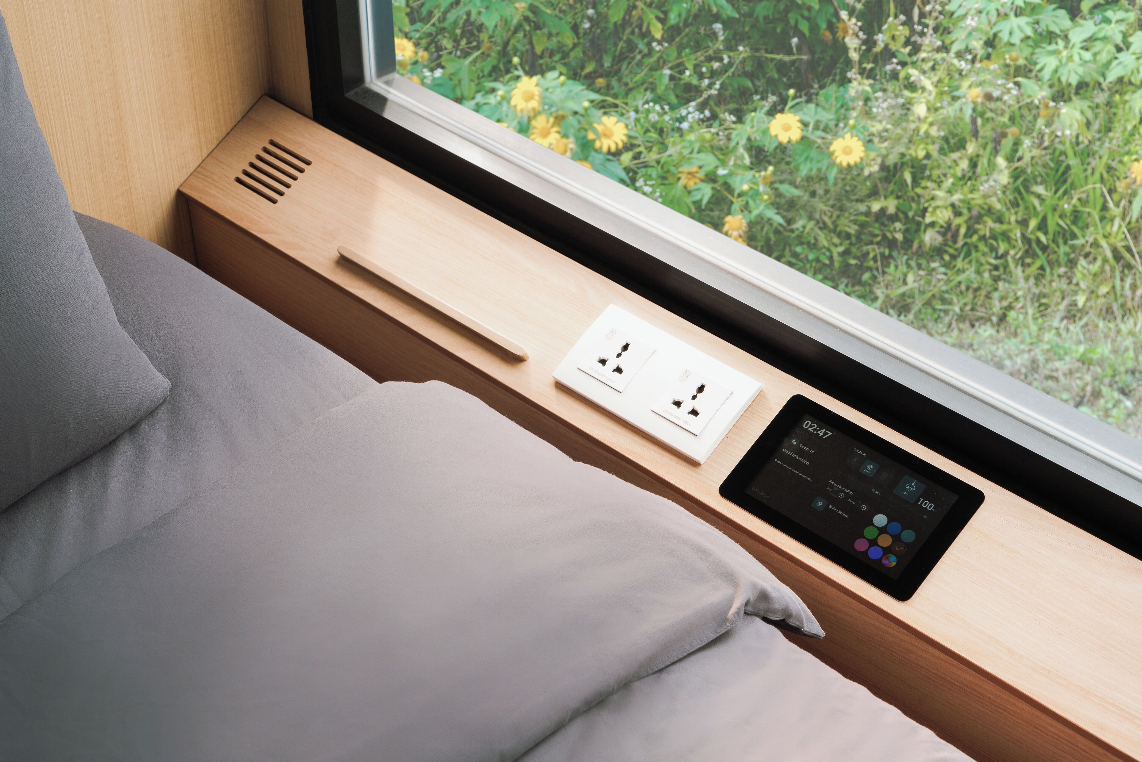 A modern room with a bed, a window with a view of greenery, a bedside table with a power outlet, and a digital device displaying time and settings.