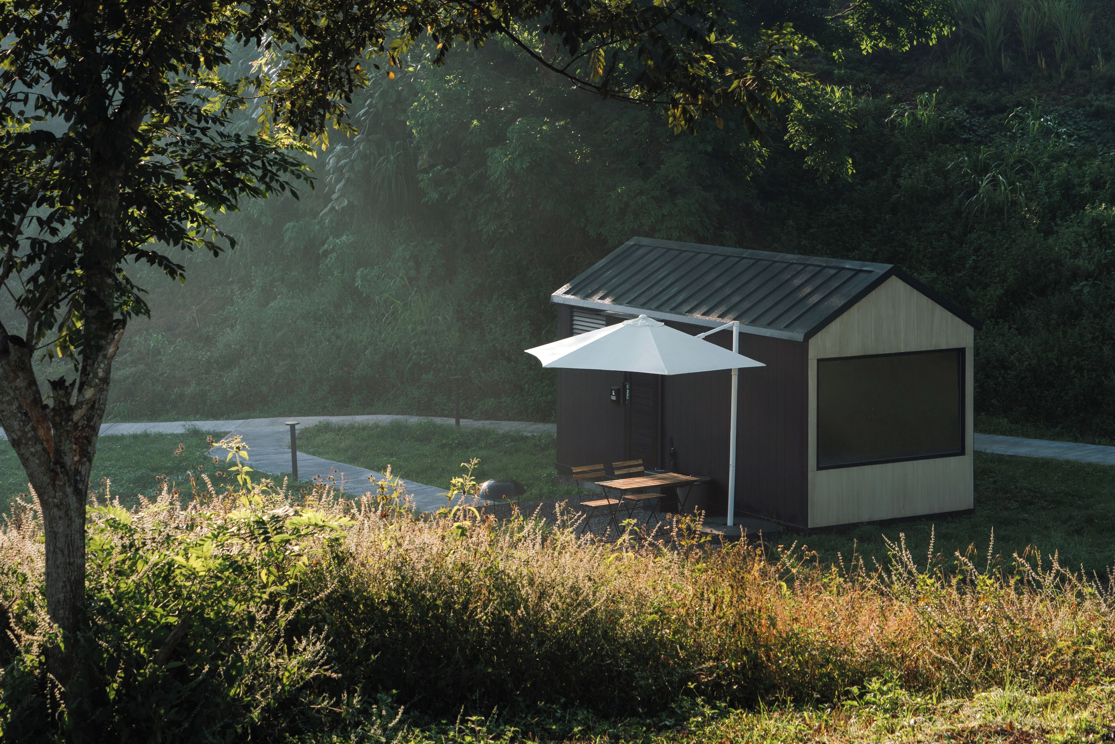 Eine kleine, moderne Holzhütte mit geneigtem Dach, einer Holzterrasse und einem weißen Sonnenschirm.