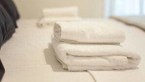 A neatly folded white towel stacked on a bed with a plain white headboard and a window in the background. - Fionia In Gazi- Athens Center Apartment Num3585 (Athens)