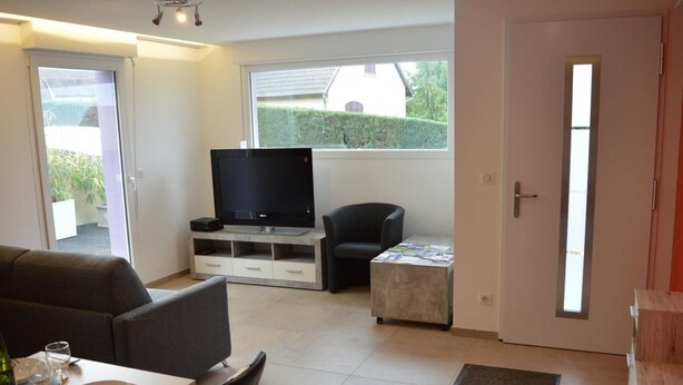 A modern living room with a flat-screen TV, a sofa, a dining table set for two, and a small coffee table. - Charming Gîte with Garden and Child's Play Area - Near Altkirch Center (Altkirch)