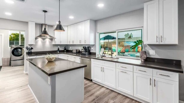 A modern kitchen with white cabinets, a central island, pendant lighting, and a view of a swimming pool. - The Monterey | Pool, Sport Court & Putting Green (Scottsdale)