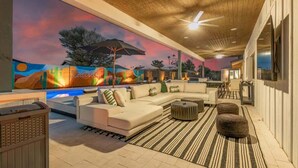 A spacious outdoor lounge area with a large sectional sofa, a round coffee table, and a striped rug. A mural with a sunset and desert landscape decorates the wall. - The Monterey | Pool, Sport Court & Putting Green (Scottsdale)