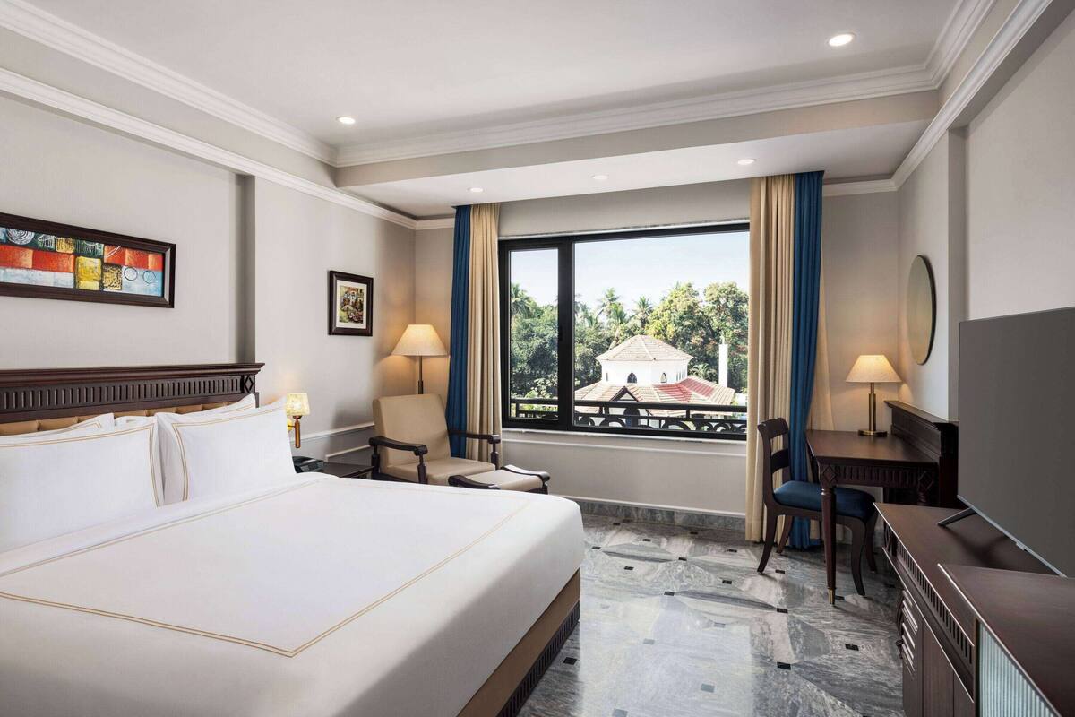 A hotel room with a large bed, a desk, two chairs, a window with a view of buildings, and artwork on the walls.