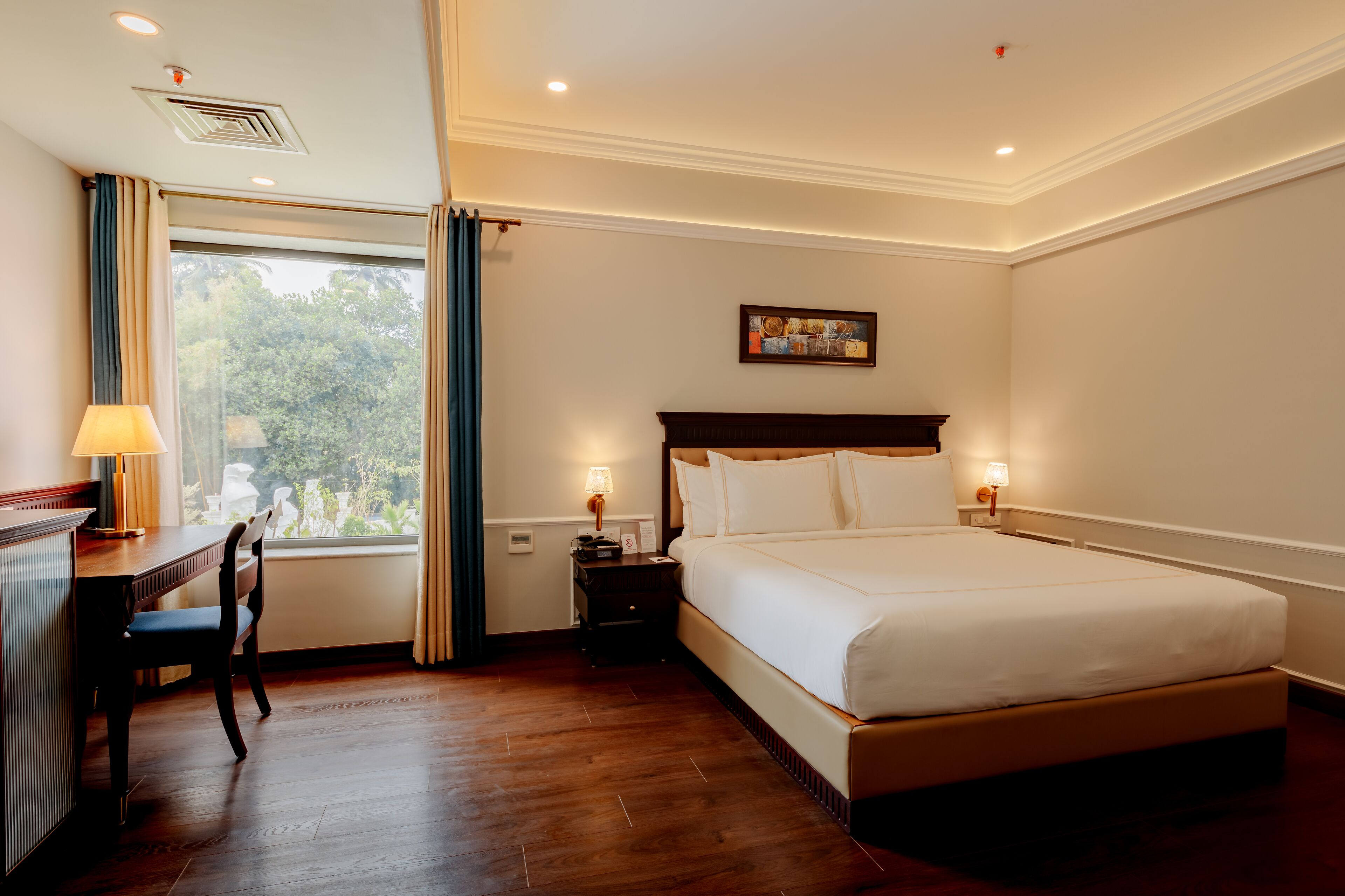 A hotel room with a large bed, a desk with a chair, a window with curtains, and a painting on the wall.