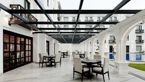 Een ruime eethoek buiten met meerdere tafels en stoelen, een zwembad en een overdekte doorgang met balkons.