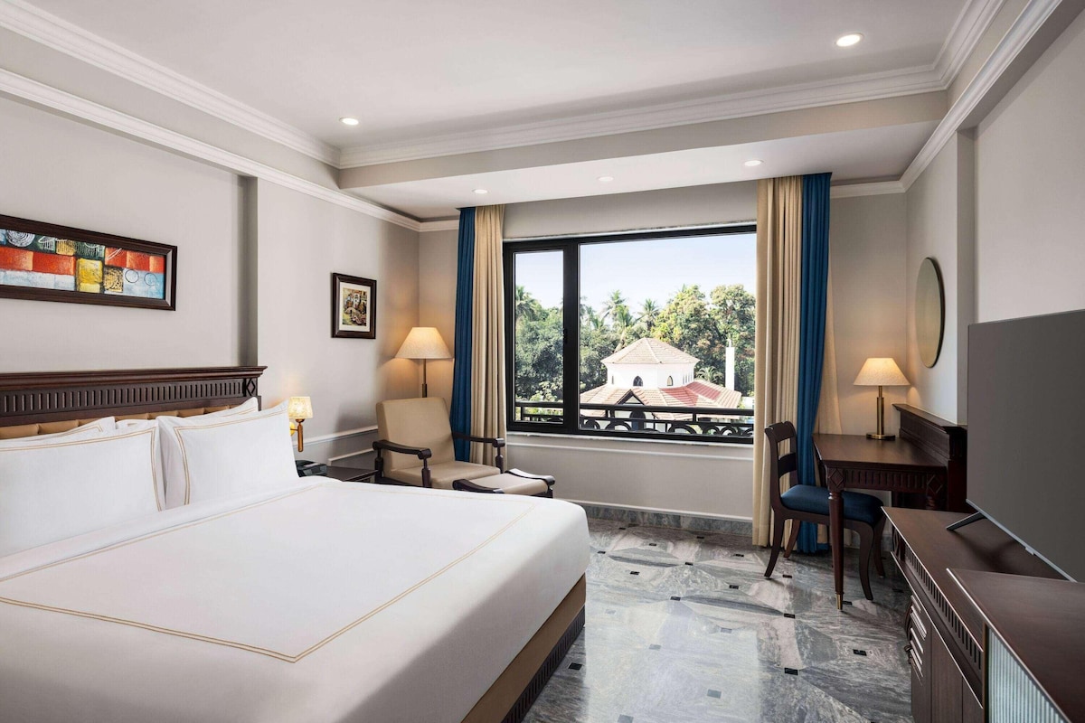 A hotel room with a large bed, a desk, a chair, a window with a view, and a painting on the wall.