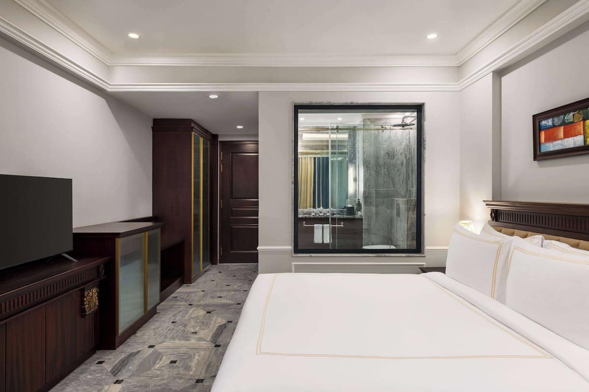 A hotel room with a large bed, a television, a wardrobe, and a bathroom visible through a glass door.