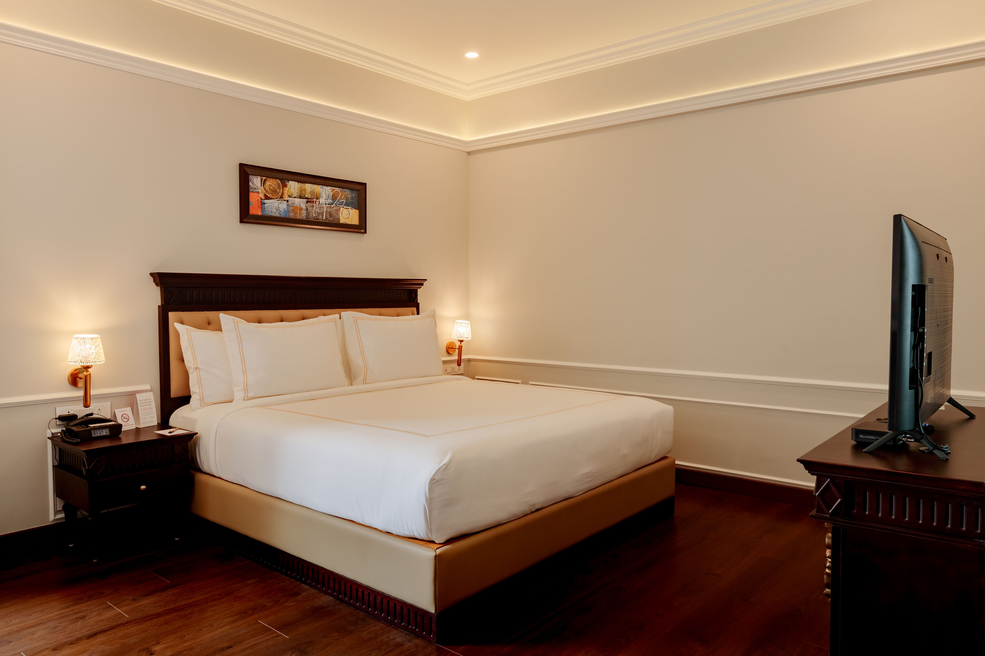 Ein Hotelzimmer mit einem großen Bett, einem hölzernen Kopfteil, zwei Nachttischlampen, einem Flachbildfernseher auf einem Holzständer und einem gerahmten Bild an der Wand.