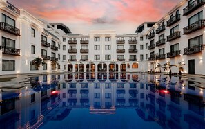 Una gran piscina interior con una fuente central, rodeada por varios pisos de un edificio blanco con balcones y superficies de piscina reflectantes.