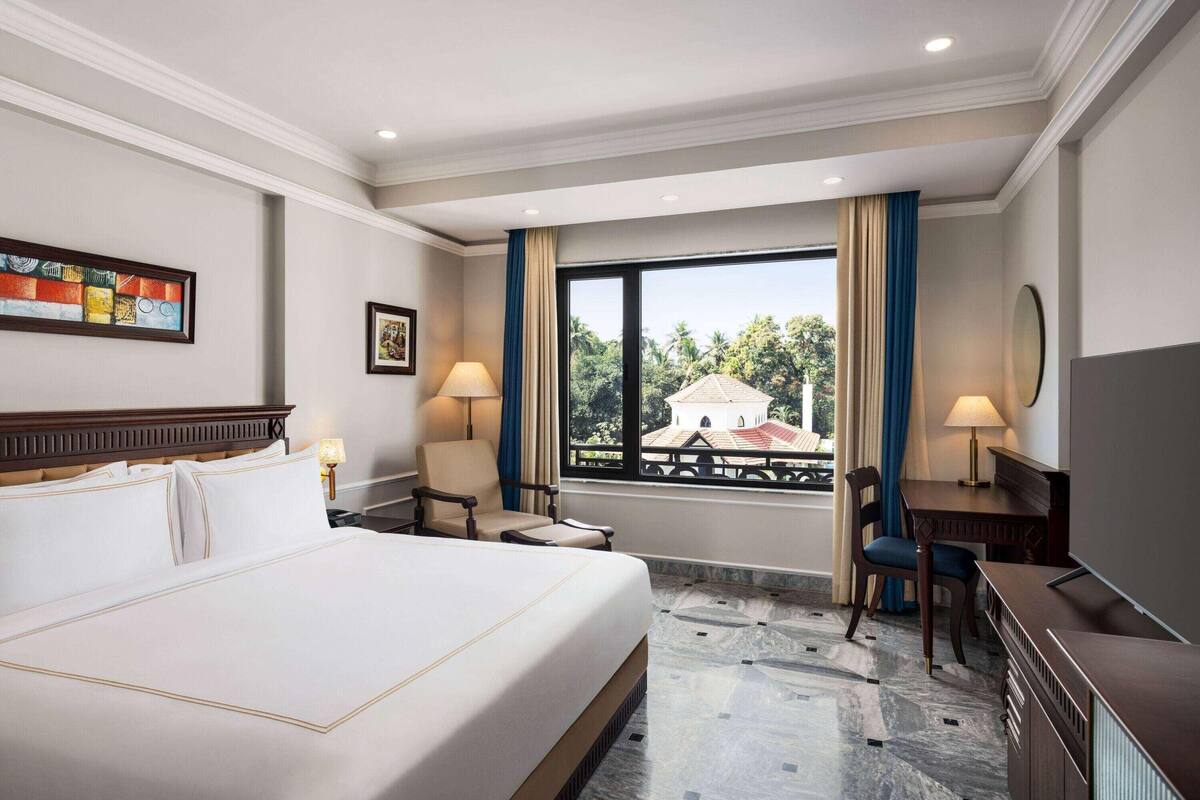 A hotel room with a large bed, a desk, a chair, a window with a view, and a painting on the wall.