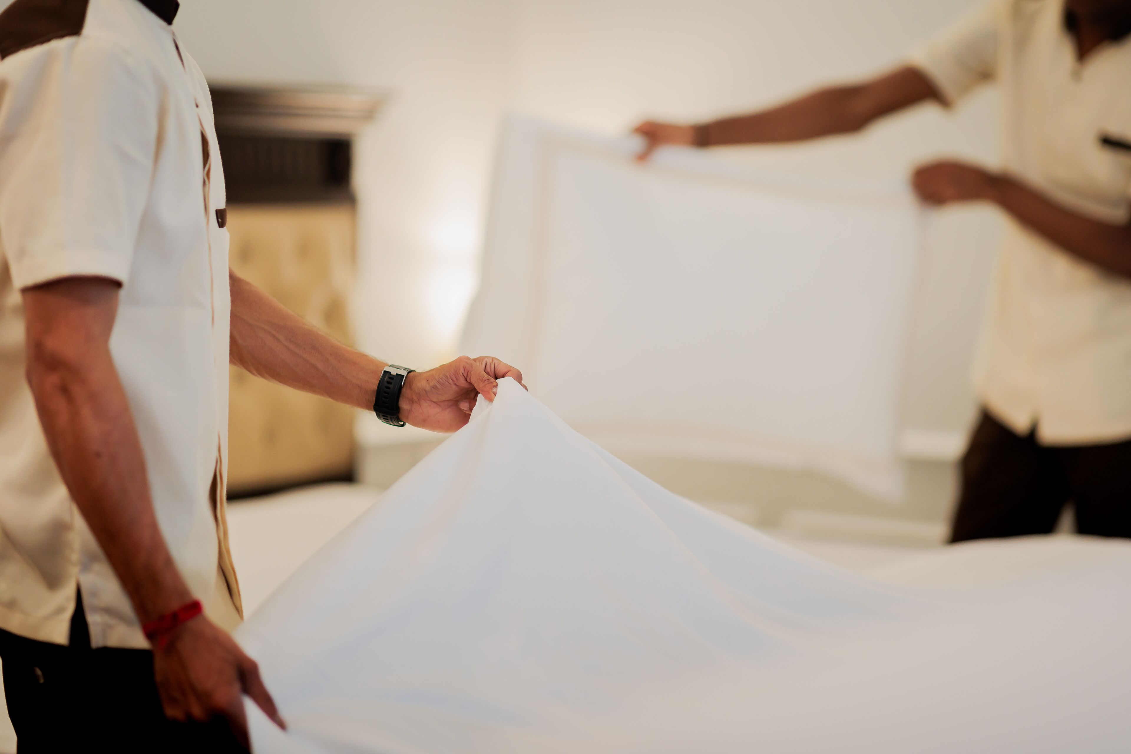 A person holding a large white sheet, possibly a bedspread, in a room with a neutral color scheme.