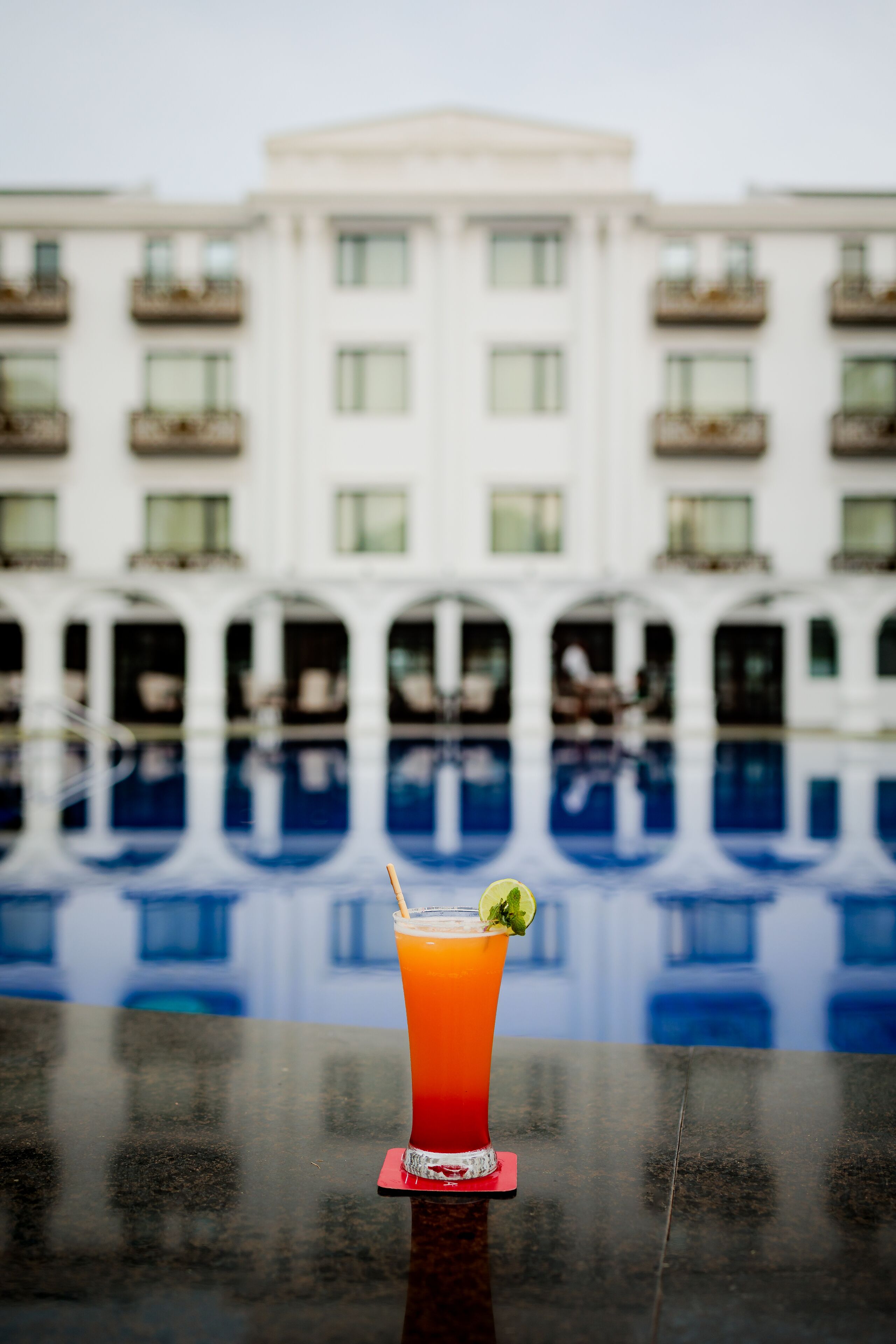 Um cocktail sobre uma superfície refletora, com um edifício e piscina ao fundo.