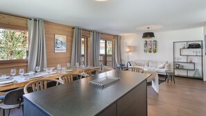 Un espace de repas moderne avec une longue table en bois dressée pour un repas, un îlot de cuisine doté d’un barbecue intégré et un coin salon confortable avec un canapé et des fauteuils.
