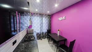 Une cuisine moderne avec un réfrigérateur noir, un coin repas avec une table et des chaises, et un mur violet sur lequel est écrit le mot « MAISON ».