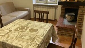 A dining area with a table covered by a patterned tablecloth, a stone fireplace, and a window with blinds. - HAPPY HOME (Orte)