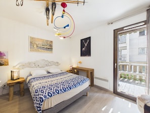 Ein Schlafzimmer mit einem Bett, einem Nachttisch, einer Lampe, einem Bild eines Rehs und einem Fenster mit Blick auf GebÀude.