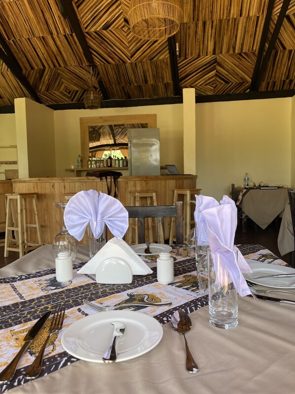 A dining area with a table set for two, featuring a patterned tablecloth, white plates, cutlery, and glassware. The ceiling has a geometric design, and there is a bar area in the background. - NJIA PANDA TENTED LODGE AND CAMPSITE (Karatu)