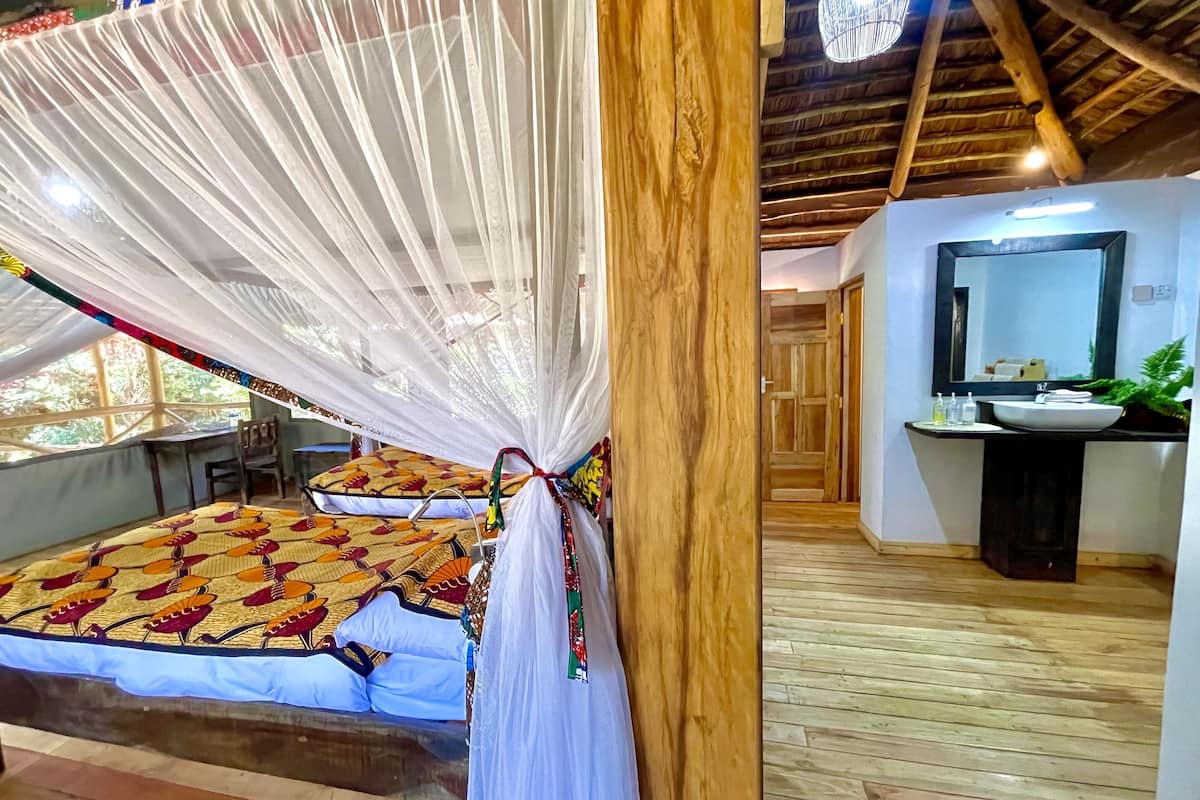 A room with a bed under a canopy, a wooden floor, a bathroom with a sink and mirror, and a desk with a chair.