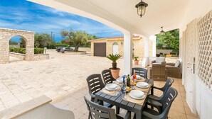 Un patio exterior con suelo de piedra, una mesa dispuesta con platos, vasos y una botella, rodeado de sillas.