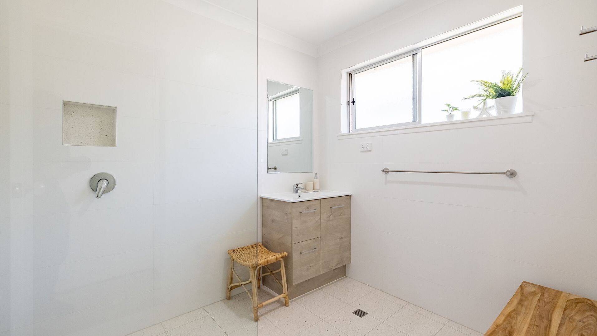 Un baño con pared blanca, tocador de madera, un taburete pequeño, una ventana con plantas y un perchero para toallas.