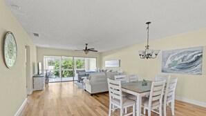 A spacious living area with a dining table, white chairs, a ceiling fan, and a large window offering a view of greenery outside.