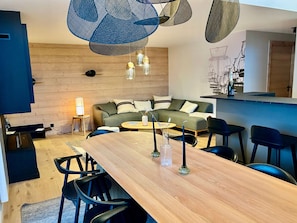 Ein modernes Wohnzimmer mit Holzboden, einer Couch, einem Esstisch mit Stühlen und hängenden Pendelleuchten.