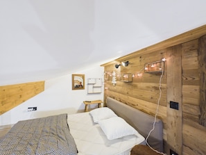 Ein Schlafzimmer mit schrägverdeckter Decke, Holzvertäfelung, einem Bett mit gemustertem Überwurf, einem Nachttisch, einer Wandablage und Lichterketten.