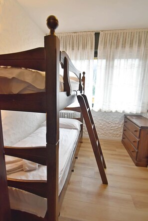 A bunk bed with a wooden headboard, a bedside table, a dresser, and a window with sheer curtains.