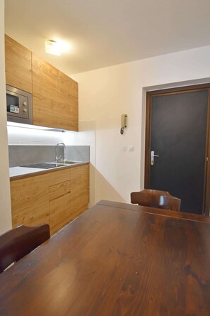 A compact kitchen with wooden cabinets, a built-in microwave, and a sink. There is a dining table with chairs and a dark door in the background.