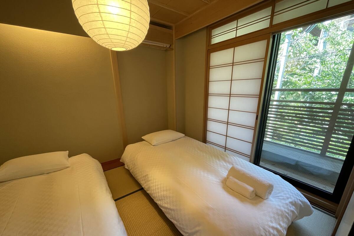 Une pièce de style japonais traditionnel, avec un sol recouvert de tatamis, deux lits avec du linge de lit blanc, un paravent shoji et une lanterne en papier suspendue.